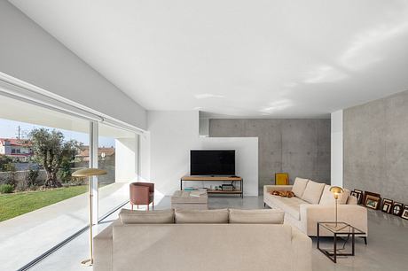 A modern, minimalist living room with concrete walls, neutral-toned furnishings, and a large window overlooking a garden.