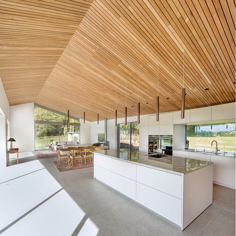 A modern kitchen with a wooden ceiling, sleek white cabinets, and a large window overlooking the outdoors.