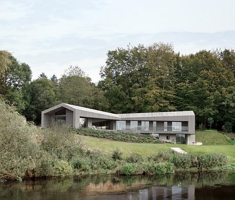 Modern, single-story home with large windows and a slanted roof, nestled in a lush, wooded landscape.