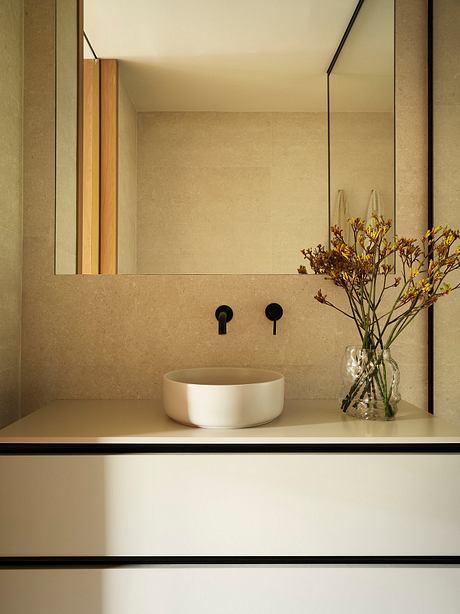 A minimalist bathroom with a white ceramic vessel sink, wooden accents, and a vase of flowers.