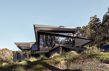 Modern glass and steel residential home perched on a sloped hillside surrounded by lush vegetation.