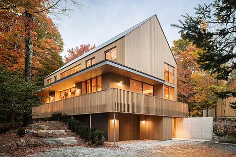 A modern wooden house with a sloped roof, balconies, and large windows nestled in an autumn forest.