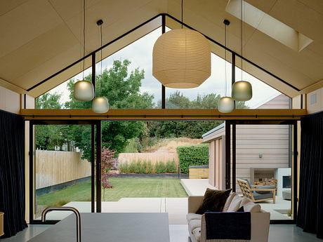 Spacious living space with vaulted ceiling, modern lighting, and panoramic glass walls overlooking lush greenery.