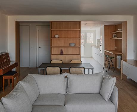 The image showcases a modern, open-concept living space with a built-in wooden bookshelf, dining table, and neutral-toned furnishings.