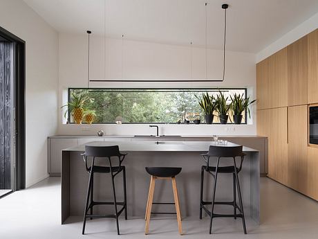Modern, minimalist kitchen with sleek, gray island and bar stools, floor-to-ceiling windows offering scenic garden views.