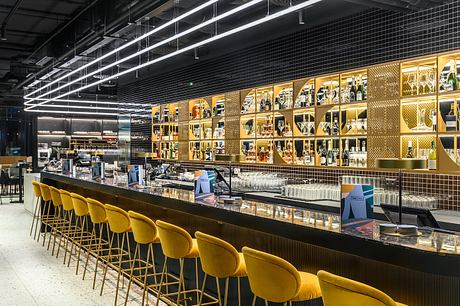 Sleek, modern bar interior with rows of yellow stools, gold-accented shelving, and dramatic lighting.