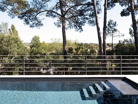 The image shows a swimming pool surrounded by a modern, minimalist architectural design with sleek metal railings and a serene forested backdrop.