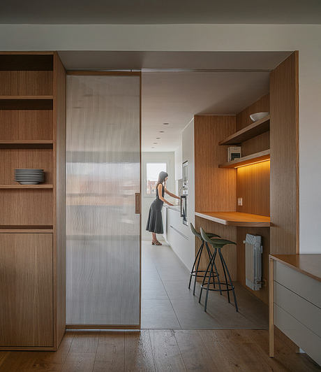 Sleek, minimalist kitchen with wood cabinetry, sliding glass partition, and modern bar stools.