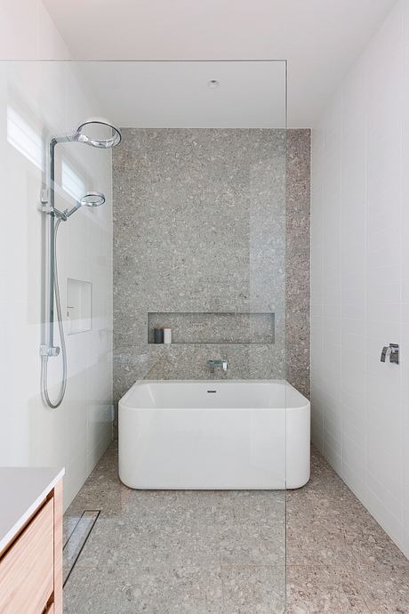 A modern bathroom with a sleek, minimalist design featuring a large white bathtub and a granite-tiled wall.