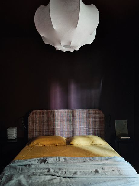 A dimly lit bedroom with a textured headboard, plush bedding, and an overhead light fixture.