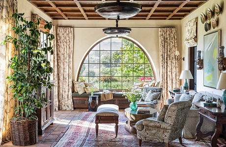 Cozy, sun-filled living room with arched window, wooden ceiling, and lush furnishings.