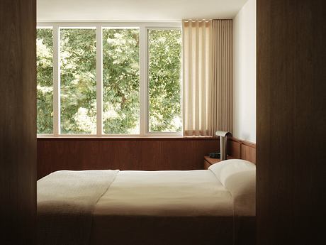 A cozy bedroom with a large window overlooking lush greenery, wooden paneling, and curtains.