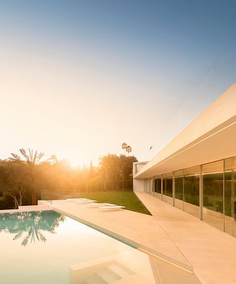 Modern glass and concrete residence with a pool, surrounded by lush greenery at sunset.