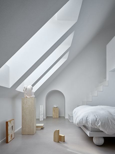 Minimalist bedroom with white walls, angled ceilings, and sculptural wooden elements.